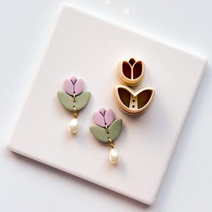 May include: A pair of handmade earrings and two polymer clay cutters. The earrings feature a lavender tulip design with a green base and a dangling pearl. The cutters are in the shape of a tulip. The items are on a white surface.