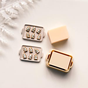 Puede incluir: Una composición festiva con galletas de jengibre en miniatura con forma de árbol en una bandeja gris, junto a un jabón rectangular y una bandeja con un inserto de jabón. Las galletas tienen detalles blancos y verdes. La escena está sobre una superficie blanca.