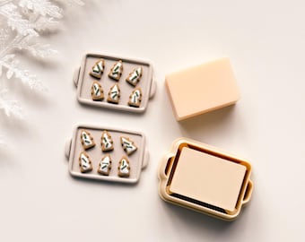 Könnte beinhalten: Eine festliche Anordnung mit Miniatur-Lebkuchenbaum-Keksen auf einem grauen Tablett, neben einem rechteckigen Seifenstück und einem Tablett mit einem Seifeneinsatz. Die Kekse haben weiße und grüne Akzente. Die Szene ist auf einer weißen Oberfläche.