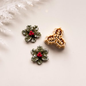 May include: Three Christmas-themed decorations. Two are green holly leaf shapes with red berries and silver glitter accents. The third is a tan-colored holly leaf shape.