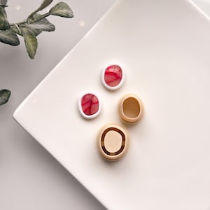 Puede incluir: Dos pendientes de resina roja y blanca con un patrón de remolino, expuestos en un plato blanco con dos respaldos de pendientes beige.