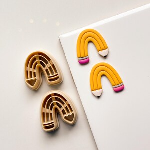 May include: An overhead shot shows two sets of rainbow-shaped objects. One set is a light brown color, and the other set is yellow with pink and white accents, resembling pencils. The objects are on a white surface.