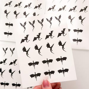 May include: Three white sheets of paper featuring multiple small, black, glittery dragon silhouettes in various flying poses. The dragons are arranged in rows and columns, showcasing different wing positions and tail shapes across the sticker sheets.