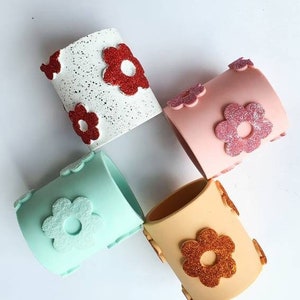 Puede incluir: Cuatro macetas decorativas de diferentes colores. Las macetas están hechas de un material texturizado y tienen un diseño de flor en la parte delantera. Los colores son blanco con flores de purpurina roja, rosa con flores de purpurina rosa, azul claro con flores de purpurina blanca y naranja con flores de purpurina naranja.