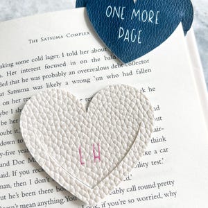 Puede incluir: Dos marcadores de página en forma de corazón. Uno es blanco con letras rosas y el otro es azul con el texto "One More Page".