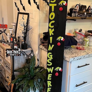 Puede incluir: Un letrero de madera negro y alto con el texto "THE STOCKINGS WERE HUNG" en letras verdes. El letrero está decorado con elementos temáticos de Grinch y adornos rojos, perfecto para Halloween o Navidad.