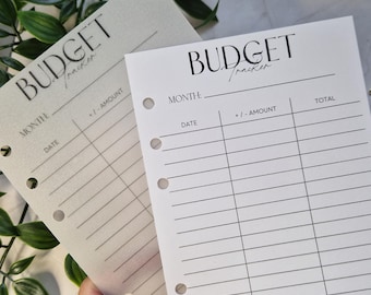 Puede incluir: Dos hojas de seguimiento presupuestario con la palabra "BUDGET" en una fuente elegante. Las hojas tienen líneas para el seguimiento de gastos, con secciones para el mes, la fecha y el importe. Las hojas se sostienen en una mano, con follaje verde en el fondo.