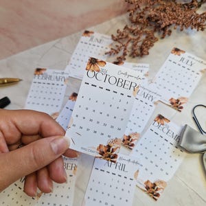 May include: A selection of small, white 2026 calendars, each showing a different month. The October calendar is held in a hand. Floral accents and the text "Create your calm" are visible. A pen and a ribbon are also in the image.
