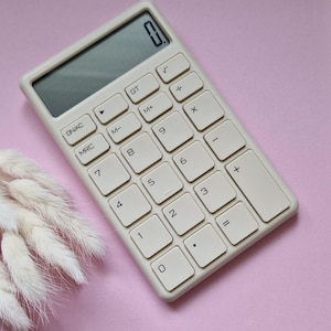 Könnte beinhalten: Ein cremefarbener Taschenrechner mit großen, quadratischen Tasten und einem Digitaldisplay mit der Anzeige "0". Der Rechner hat ein minimalistisches Design. Die Tasten umfassen Zahlen, grundlegende mathematische Symbole und Funktionstasten.