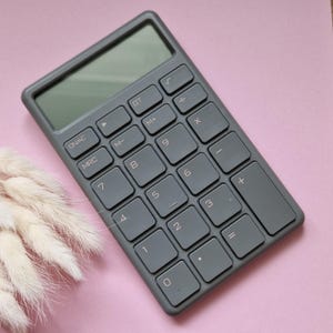 Peut inclure: Une calculatrice grise de forme rectangulaire avec un écran numérique. La calculatrice a des boutons carrés avec des chiffres et des symboles blancs. La calculatrice est sur une surface rose.