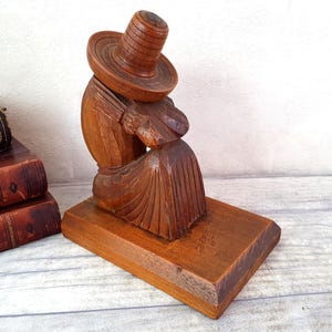 Peut inclure: Un serre-livres en bois sculpté représentant une personne portant un chapeau à larges bords. La figure est assise et semble tenir quelque chose sur ses genoux. Le serre-livres est en bois foncé et a une finition naturelle.