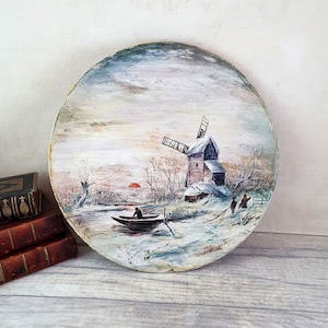 May include: A round wooden plate featuring a painted scene of a windmill in a snowy landscape. The windmill is white with a red roof and is surrounded by trees and a frozen river. Two figures are depicted in the foreground, one in a boat and the other on the shore. The sky is a pale blue with a hint of pink.