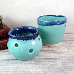 May include: Two ceramic planters in a light blue-green glaze with a dark blue rim. The smaller planter has drainage holes.