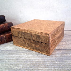 Puede incluir: Una caja rectangular de madera con tapa con bisagras. La caja es de madera marrón claro con vetas visibles y un acabado natural. La caja está cerrada y se encuentra sobre una superficie gris claro. Libros antiguos en el fondo.