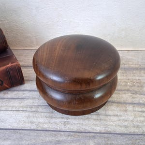 Op de afbeelding: Een donkerbruine houten hoedenblok met een afgeronde boven- en onderkant. Het hoedenblok is gemaakt van massief hout en heeft een gladde, gepolijste afwerking. Het is ontworpen voor het vormen van hoeden en is een handig hulpmiddel voor de hoedenmakerij.