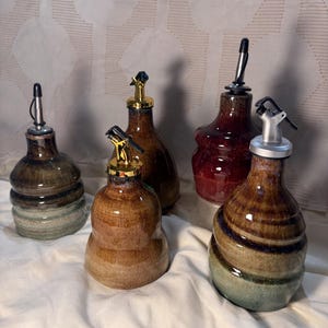 May include: Five ceramic oil dispensers with glossy glazes in shades of brown, green, and red. Each bottle features a rounded base, a tapered neck, and a metal pour spout with a hinged cap. The glazes show variations in color and texture.