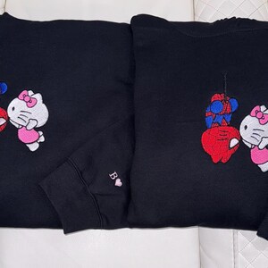 Pu&ograve; includere: Due felpe nere con disegni ricamati. Uno presenta un Spider-Man rosso e blu e una Hello Kitty bianca con un vestito rosa. L'altro ha lo stesso design. Entrambe le maniche hanno una lettera e un cuore.