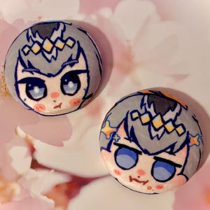 May include: Two round, plush buttons with anime-style character faces. The characters have gray hair, blue eyes, and pink cheeks. The background is a soft pink with white flower petals.