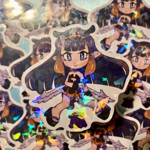 May include: A sticker featuring a cartoon character with purple hair, a crown, and wings. The character is wearing a black and white outfit and has a halo. The sticker has a holographic finish.