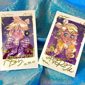 May include: Two polaroid-style prints featuring anime-style illustrations of a character with blonde hair and blue eyes. The prints have gold foil accents and handwritten text. The background is a shimmering blue fabric.