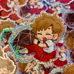 May include: A holographic sticker featuring a cartoon character with brown hair and glasses, wearing a white shirt with red trim and red pants, hugging a large, dark brown, multi-faceted geometric shape resembling a twenty-sided gaming die.