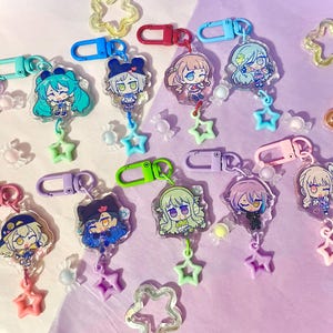 Puede incluir: Una colección de llaveros de personajes de estilo anime con detalles en forma de estrella y plástico transparente. Cada llavero presenta un diseño de personaje diferente, un cierre de color y un dije en forma de estrella. El fondo es de color lila claro.