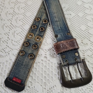 May include: A blue denim belt featuring two rows of brass-toned metal grommets, a dark brown leather loop, and a large, weathered brass-toned double-prong buckle. The belt end is finished with a black leather tip and a small red rectangular tag.
