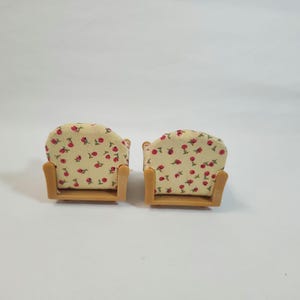 May include: Two miniature armchairs. The chairs feature a light beige fabric with a red floral pattern. The frames are made of light brown wood. The chairs are designed for a doll's house or miniature setting.
