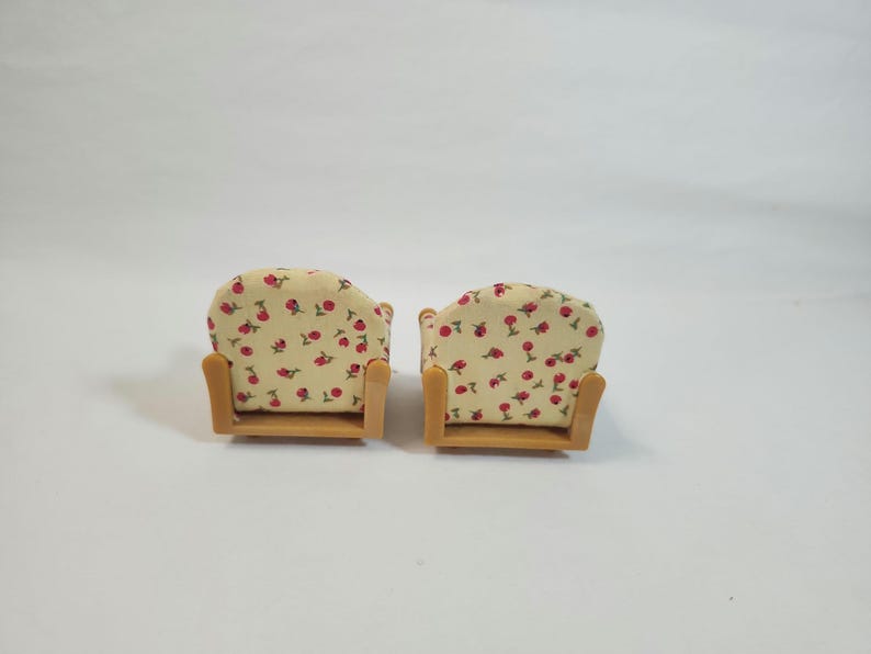 May include: Two miniature armchairs. The chairs feature a light beige fabric with a red floral pattern. The frames are made of light brown wood. The chairs are designed for a doll's house or miniature setting.