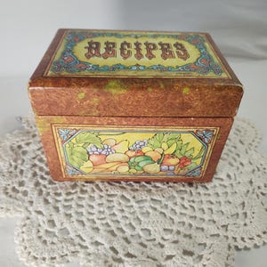 May include: A rectangular brown recipe box with a mottled finish. The lid features the word RECIPES in stylized lettering. The front panel displays a colorful illustration of various fruits and vegetables, including grapes, pears, and carrots.
