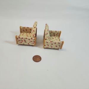 May include: Two miniature armchairs with a light beige fabric covered in red cherry patterns. The wooden frames have a natural finish. A penny is next to the chairs for scale. The chairs are likely for a dollhouse or miniature setting.