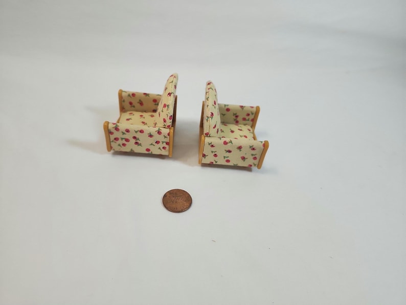 May include: Two miniature armchairs with a light beige fabric covered in red cherry patterns. The wooden frames have a natural finish. A penny is next to the chairs for scale. The chairs are likely for a dollhouse or miniature setting.