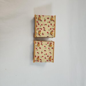 May include: Two small, square wooden wall hooks. Each hook has a light yellow fabric surface with a red cherry pattern. The wooden frames are light brown, and each hook has four round, light brown knobs.