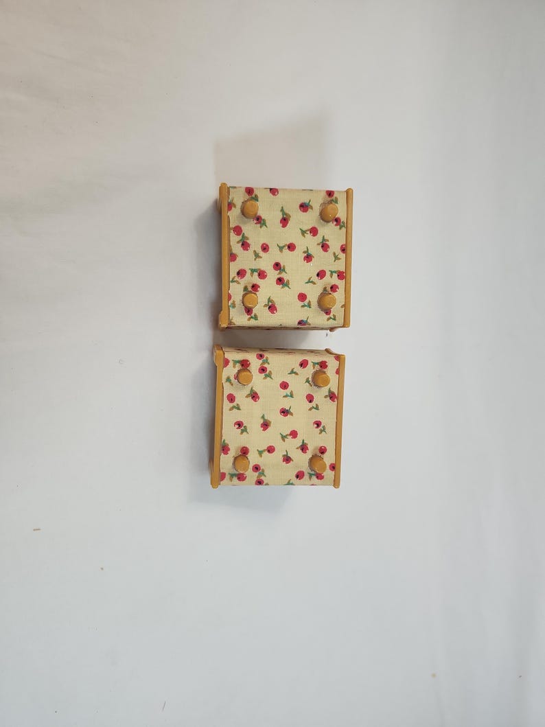 May include: Two small, square wooden wall hooks. Each hook has a light yellow fabric surface with a red cherry pattern. The wooden frames are light brown, and each hook has four round, light brown knobs.