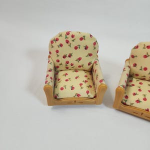 May include: Two miniature armchairs. The chairs feature a light beige fabric adorned with a pattern of red flowers and green leaves. The frames are a light brown colour. These chairs are designed for a dollhouse or miniature setting.