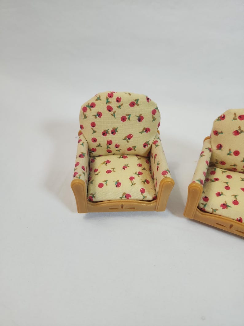 May include: Two miniature armchairs. The chairs feature a light beige fabric adorned with a pattern of red flowers and green leaves. The frames are a light brown colour. These chairs are designed for a dollhouse or miniature setting.