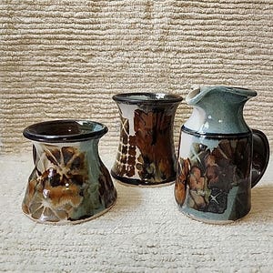 May include: Three handcrafted ceramic vessels with a rustic aesthetic. The pieces feature a mix of brown, green, and white glazes, with leaf-like patterns. One is a pitcher with a handle, and the others are small vases.