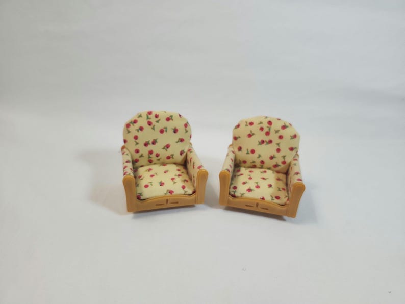 May include: Two miniature armchairs. The chairs feature a light beige fabric patterned with red cherries and green leaves. The frames are light brown. The chairs are set against a white background.