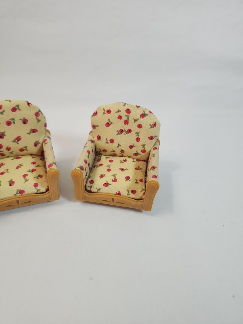 May include: Two miniature armchairs. The chairs feature a light yellow fabric with a red cherry pattern. The armrests and frame are light brown. These chairs are designed for a doll's house or similar miniature setting.