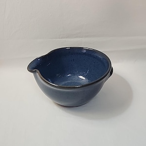 May include: A handmade, blue ceramic bowl with a spout and small handle. The bowl has a speckled texture and a dark rim. The interior is a glossy, deep blue. This unique bowl is perfect for serving sauces or dips.