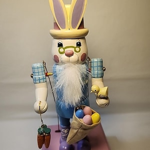 Puede incluir: Un cascanueces de madera con forma de conejito de Pascua, con cara blanca, barba larga blanca y gafas redondas. El conejito viste un traje azul y un sombrero con orejas moradas. Sostiene una bolsa de huevos de colores y una zanahoria. Un pollito amarillo está en su brazo.