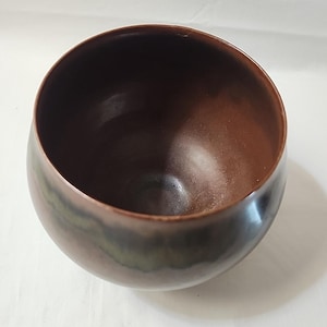 以下が含まれることがあります： 手作りの丸いボウルで、内側は濃い茶色、外側はグラデーションになっています。外側は濃い、ほぼ黒に近い色から緑色に変化し、ユニークな視覚効果を生み出しています。ボウルは滑らかで磨かれた表面をしています。