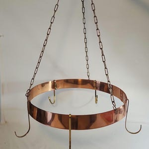 May include: A circular copper pot rack featuring a wide, polished copper band suspended by three metal chains. The rack includes several brass-colored hooks attached to the rim, designed for hanging kitchen items from the frame.