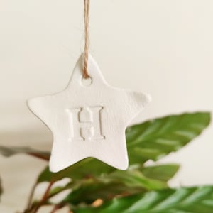 May include: White clay star ornament with the letter "H" embossed on it. The star is hanging from a twine string.