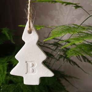 May include: White ceramic Christmas tree bauble with the letter "B" embossed on the front. The bauble is hanging from a twine string.