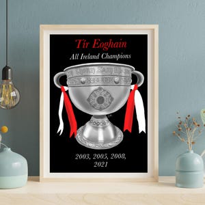 Może przedstawiać: Oprawiony wydruk przedstawiający srebrny puchar na czarnym tle. Tekst zawiera napis Tír Eoghain, All Ireland Champions oraz lata 2003, 2005, 2008, 2021. Czerwone i białe wstążki są przywiązane do uchwytów zilustrowanego pucharu.