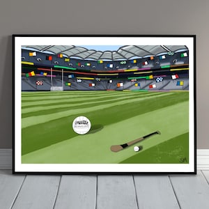 Może przedstawiać: Oprawiona cyfrowa ilustracja boiska sportowego z białą piłką do futbolu gaelickiego O'Neills, drewnianym kijem do hurlingu i małą białą piłeczką sliotar na trawie, z kolorowymi flagami zawieszonymi na trybunach stadionu.