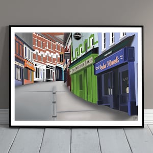 Może przedstawiać: Oprawiona ilustracja cyfrowa przedstawiająca scenę uliczną z witrynami sklepowymi z szyldami The Fweedore Bar i Peador O'Donnell's. Budynki są utrzymane w kolorach czerwonym, zielonym, niebieskim i białym, wzdłuż szarej ulicy ze słupkiem.