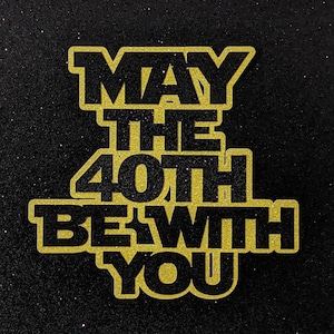May include: A black and gold glitter cake topper with the text "MAY THE 40TH BE WITH YOU". The text is in a bold, blocky font, with a gold glitter outline and black fill. The background is black glitter.