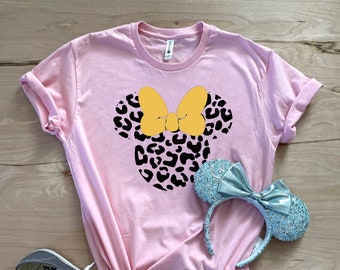 May include: Pink t-shirt with a black and white leopard print Minnie Mouse head design with a yellow bow. The shirt is laying on a wooden surface with a pair of gray Vans shoes and a blue glitter Minnie Mouse headband.